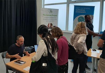 Image of people networking at Pathways to Employment Event. Two signs in the background with the text: The Autism Project and West Lea School Supported Internships 