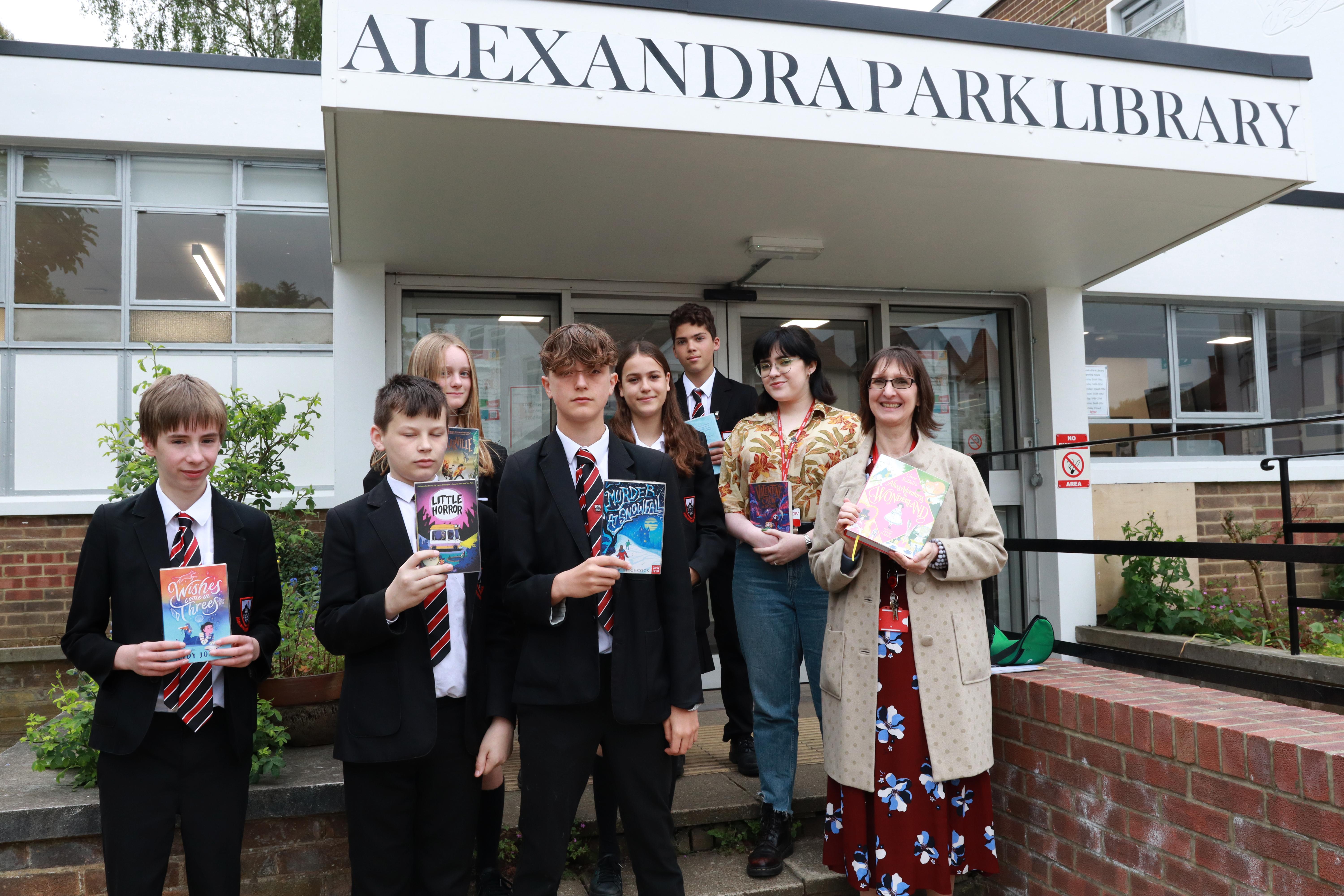 The picture is of six Alexandra Park School (APS) pupils and two Haringey Libraries staff stood outside Alexandra Park Library, with each of them holding up a book from the library.